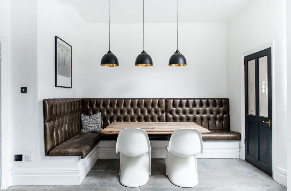 A stylish dining nook featuring a tufted leather banquette, rustic wooden table, sleek white chairs, and black pendant lights, showcasing a perfect blend of modern and classic commercial furniture design.